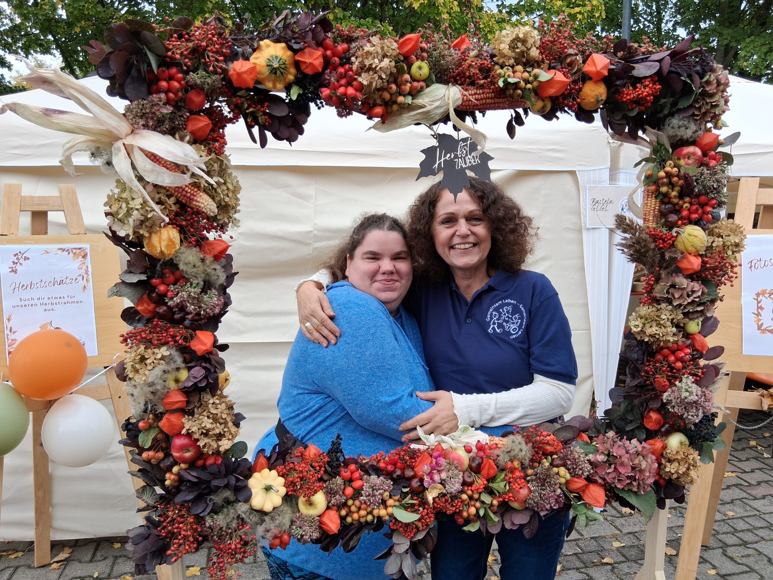 1000002037 Foto von zwei Frauen (eine Betreuerin und eine Klientin), die sich im Arm halten und in die Kamera lächeln. Sie stehen hinter einem herbstlich dekorierten Rahmen.